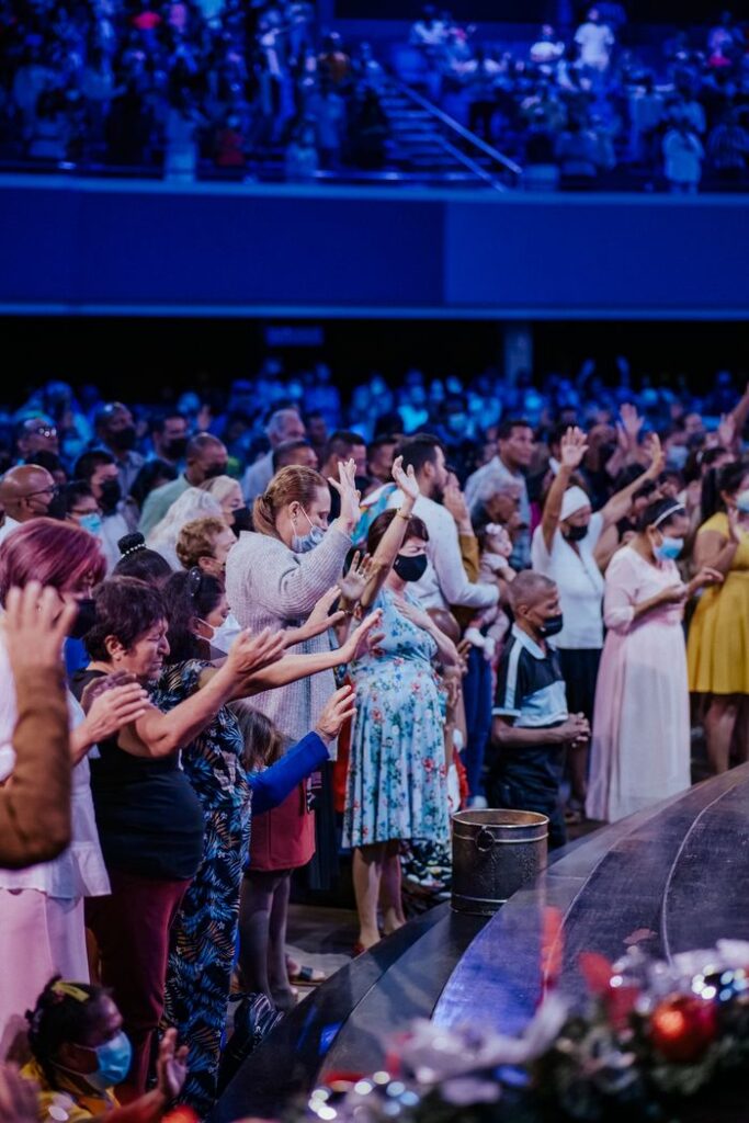 A large group of diverse people standing with arms raised, showcasing unity and diversity indoors.